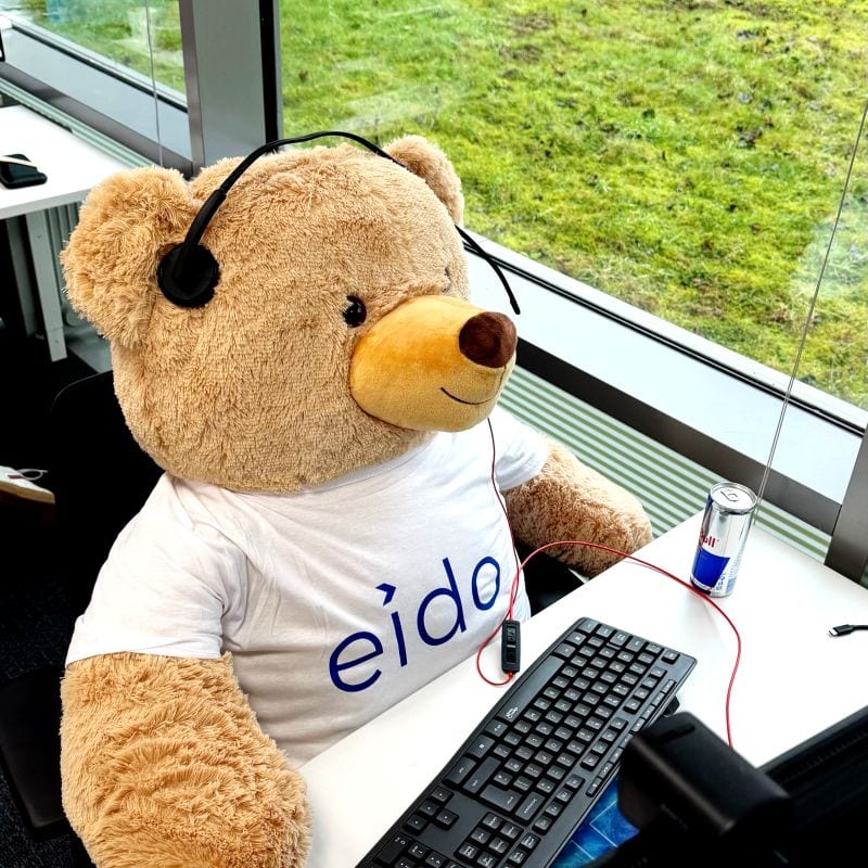 Odie, a large teddy bear in an Eido T-shirt, sitting at a desk wearing a headset.