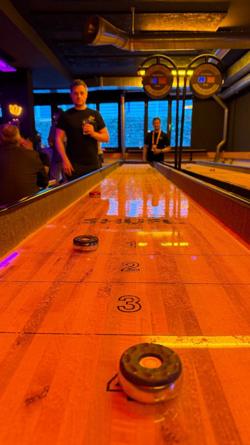 A shuffleboard table lit in orange, with pucks in the foreground and two players at the far end.